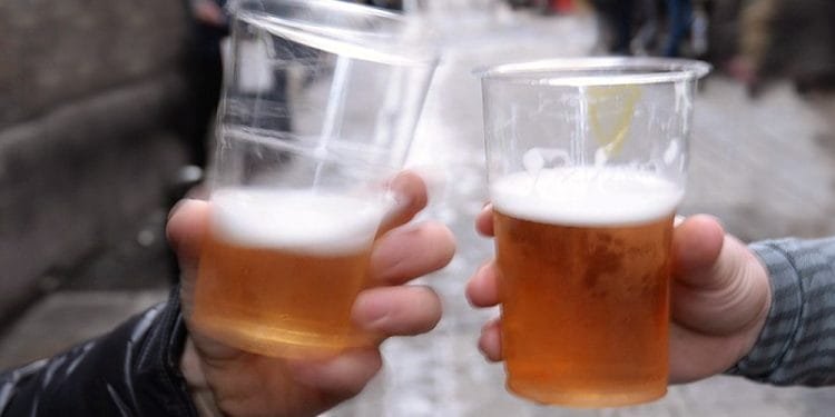 Due persone tintinnano bicchieri di plastica di birra in un ambiente esterno. Lo sfondo mostra una scena di strada sfocata con diverse persone ed edifici, dando la sensazione di un luogo pubblico e vivace, forse a Lisbona.
