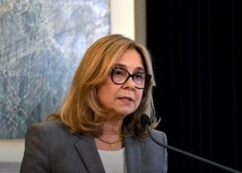 Una donna dai capelli biondi, con gli occhiali e un blazer grigio, parla al microfono. Ha un'espressione seria e sta di fronte a un dipinto astratto geometrico, discutendo del ruolo di un medico famiglia in Portogallo.