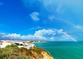 Una vista panoramica della costa in Meteo Portogallo presenta un vibrante doppio arcobaleno che si estende sulle acque turchesi dell'oceano. Case bianche con tetti di tegole rosse sono arroccate su una scogliera sotto un cielo azzurro con nuvole sparse. La vegetazione verde delimita il bordo della scogliera.