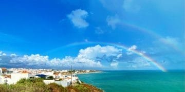 Una vista panoramica della costa in Meteo Portogallo presenta un vibrante doppio arcobaleno che si estende sulle acque turchesi dell'oceano. Case bianche con tetti di tegole rosse sono arroccate su una scogliera sotto un cielo azzurro con nuvole sparse. La vegetazione verde delimita il bordo della scogliera.