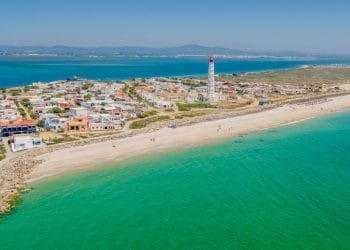 Veduta aerea di una città costiera dell'Algarve con edifici colorati e un alto faro vicino a una lunga spiaggia caraibica. L'acqua turchese dell'oceano incontra la riva in una giornata limpida e soleggiata, con le montagne visibili in lontananza.