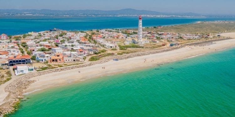 Veduta aerea di una città costiera dell'Algarve con edifici colorati e un alto faro vicino a una lunga spiaggia caraibica. L'acqua turchese dell'oceano incontra la riva in una giornata limpida e soleggiata, con le montagne visibili in lontananza.