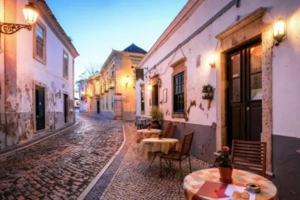 La quarta delle 10 cose da vedere in Algarve sono le strada acciottolate di Faro al tramonto, fiancheggiata da vecchi edifici imbiancati con luci calde e luminose. Piccoli tavoli di legno con sedie e piante in vaso siedono all'esterno di un bar, creando un'atmosfera invitante e accogliente.