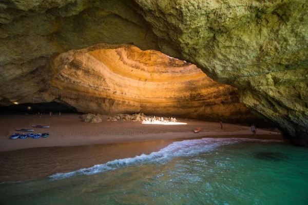 La prima delle 10 da vedere in Algarve, La grotta di Benagil con un'apertura circolare nella parte superiore che permette alla luce del sole di entrare. La grotta è circondata da acqua verde smeraldo, con alcune piccole imbarcazioni e alcune persone visibili sulla sabbia e nella grotta.