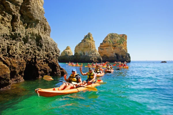 Una delle 10 cose da fare in Algarve, un gruppo di persone in kayak su acque cristalline vicino a grandi formazioni rocciose di Ponta da Piedade in Algarve. I canoisti indossano giubbotti di salvataggio e caschi e il cielo sopra è limpido e luminoso.