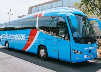 Un grande e moderno autobus blu con la scritta Rede Expressos scritta sulla fiancata è parcheggiato in una strada. L'autobus ha un design elegante con strisce rosse e bianche che vanno dal tetto fino al centro. Sullo sfondo sono visibili un albero e un edificio.