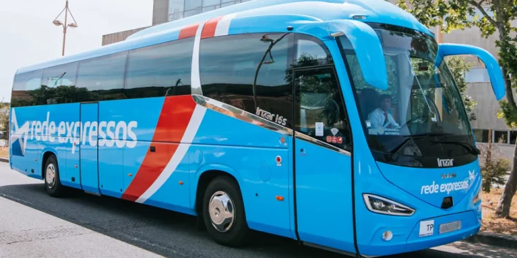 Un grande e moderno autobus blu con la scritta Rede Expressos scritta sulla fiancata è parcheggiato in una strada. L'autobus ha un design elegante con strisce rosse e bianche che vanno dal tetto fino al centro. Sullo sfondo sono visibili un albero e un edificio.