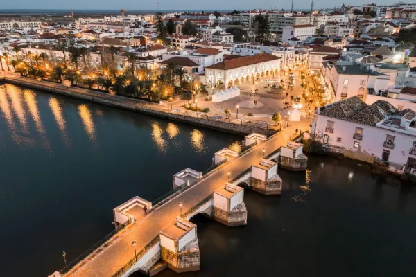 Veduta aerea serale di Tavira in Algarve, con edifici bianchi raggruppati attorno a una piazza illuminata e alberata vicino al lungomare. Un ponte romano con archi ben illuminati si estende sull'acqua serena, collegando diverse parti di questa scena idilliaca, perfetta per la tua lista delle 10 migliori cose da vedere.