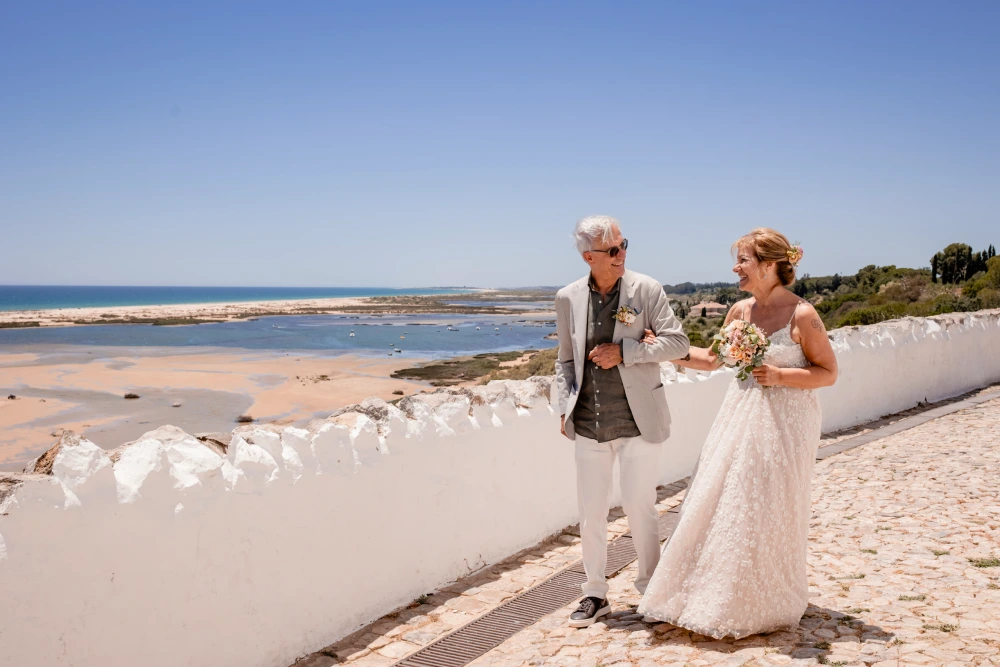 Umberto Agnese e Camilla Salerno in abiti da sposi. La sposa tiene in mano un mazzo di fiori mentre cammina a braccetto con lo sposo in abito grigio chiaro e occhiali da sole. Stanno camminando lungo un sentiero di ciottoli bianchi con una vista panoramica sulla costa dell'Algarve in Portogallo.