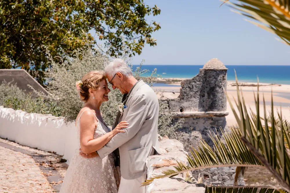 Umberto Agnese e Camilla Salerno finalmente sposi. Sorridendo sotto un albero con l'oceano sullo sfondo. Sono su un sentiero acciottolato accanto a un muro bianco, vicino a un'antica struttura in pietra, con verde e foglie di palma che incorniciano la scena.