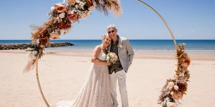Umberto Agnese e Camilla Salerno in abiti da sposi sorridenti sotto un arco floreale circolare su una spiaggia in Portogallo. La donna indossa un abito da sposa di pizzo bianco e tiene in mano un bouquet, mentre l'uomo indossa un abito leggero con occhiali da sole. L'oceano e il cielo azzurro dell'Algarve formano lo sfondo sereno.