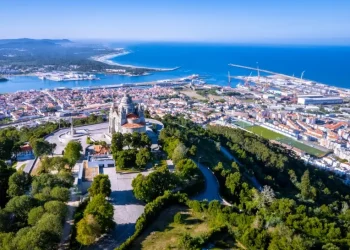 Veduta aerea di Viana do Castelo la terza città più accogliente d'Europa. Si vede il Santuário Diocesano do Sagrado Coração de Jesus circondata da alberi in cima a una collina. Il paesaggio urbano comprende un fiume, aree residenziali e un porto industriale con gru. Lo sfondo mostra la costa che incontra l'oceano blu sotto un cielo limpido.