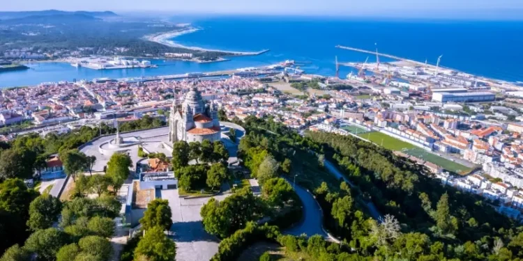 Veduta aerea di Viana do Castelo la terza città più accogliente d'Europa. Si vede il Santuário Diocesano do Sagrado Coração de Jesus circondata da alberi in cima a una collina. Il paesaggio urbano comprende un fiume, aree residenziali e un porto industriale con gru. Lo sfondo mostra la costa che incontra l'oceano blu sotto un cielo limpido.
