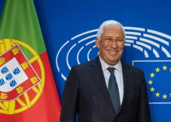 Antonio Costa, eletto Presidente del Consiglio Europeo sorride davanti a due bandiere: quella portoghese a sinistra e quella dell'Unione Europea a destra. Lo sfondo è un muro blu con linee circolari bianche. È un uomo in giacca e cravatta