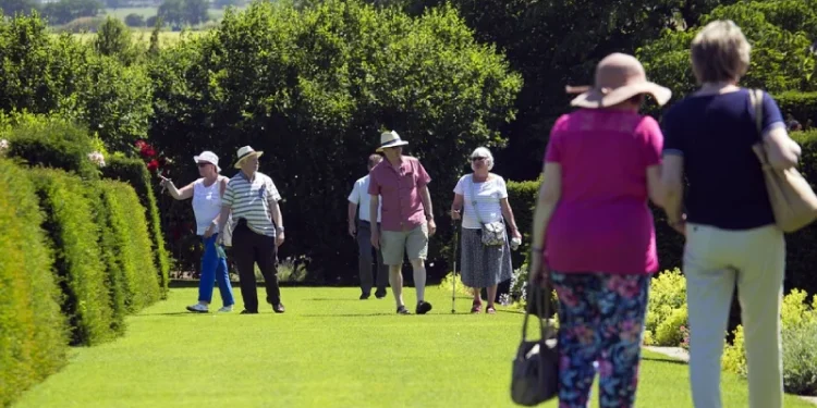 Un gruppo di anziani passeggia in un giardino verde e soleggiato in Portogallo. Alcuni indossano cappelli e una donna in primo piano indossa un cappello a tesa larga e pantaloni a fiori. Il giardino ha siepi e alberi ben curati, offrendo un'atmosfera scenografica e tranquilla sotto un cielo terso.