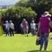 Un gruppo di anziani passeggia in un giardino verde e soleggiato in Portogallo. Alcuni indossano cappelli e una donna in primo piano indossa un cappello a tesa larga e pantaloni a fiori. Il giardino ha siepi e alberi ben curati, offrendo un'atmosfera scenografica e tranquilla sotto un cielo terso.