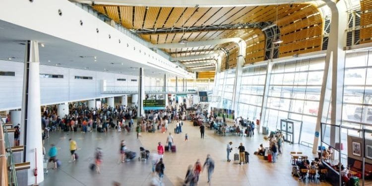 Un terminal aeroportuale affollato, con viaggiatori che camminano e aspettano in fila. Il soffitto è caratterizzato da doghe di legno e le grandi finestre offrono luce naturale. Gli schermi digitali mostrano le informazioni sui voli, compresi i consigli utili per chi ha un volo per il Portogallo.