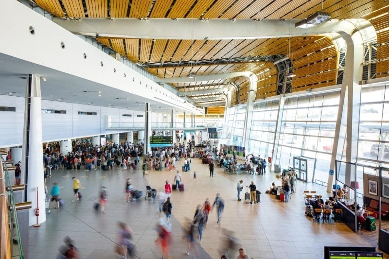 Un terminal aeroportuale affollato, con viaggiatori che camminano e aspettano in fila. Il soffitto è caratterizzato da doghe di legno e le grandi finestre offrono luce naturale. Gli schermi digitali mostrano le informazioni sui voli, compresi i consigli utili per chi ha un volo per il Portogallo.