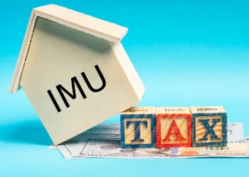 Su una pila di banconote da un dollaro si erge una casetta di legno con la scritta Esenzione IMU su un lato e fa riferimento all'estero aire. Davanti alla casa, tre blocchi colorati compongono la scritta "TAX" su uno sfondo blu.