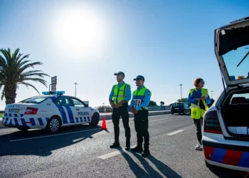 Due agenti di polizia stanno accanto a un'auto di pattuglia durante i controlli per i falsi tassisti negli aeroporti in portogallo. Un'altra macchina della polizia con la scritta policia è parcheggiata. Una terza persona, che indossa un giubbotto ad alta visibilità, tiene in mano un blocco per appunti.