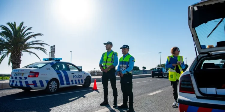 Due agenti di polizia stanno accanto a un'auto di pattuglia durante i controlli per i falsi tassisti negli aeroporti in portogallo. Un'altra macchina della polizia con la scritta policia è parcheggiata. Una terza persona, che indossa un giubbotto ad alta visibilità, tiene in mano un blocco per appunti.