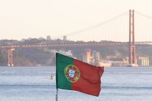 Durante la giornata nazionale del portogallo una bandiera portoghese sventola in primo piano con il ponte 25 aprile di Lisbona sullo sfondo sul fiume Tejo. Lo scenario include strutture sfocate sulla sponda opposta.