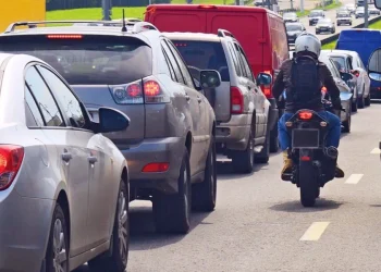 Una moto superare le code su una strada in Portogallo tra due corsie di traffico congestionate. Un motociclista con indosso un casco e uno zaino. Sono visibili diverse auto, furgoni e un SUV, con le luci dei freni accese, il che suggerisce un ingorgo. La scena sembra essere su una strada a più corsie durante il giorno.