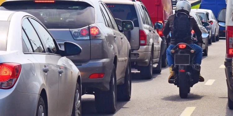 Una moto superare le code su una strada in Portogallo tra due corsie di traffico congestionate. Un motociclista con indosso un casco e uno zaino. Sono visibili diverse auto, furgoni e un SUV, con le luci dei freni accese, il che suggerisce un ingorgo. La scena sembra essere su una strada a più corsie durante il giorno.