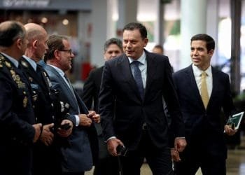 Un gruppo di uomini in abiti formali, compresi abiti e uniformi militari, camminano insieme e conversano sul nuovo piano immigrazione portogallo. Alcuni hanno in mano documenti e sembrano trovarsi in un ambiente professionale o ufficiale. Lo sfondo mostra un ambiente interno con persone e un arredamento moderno.