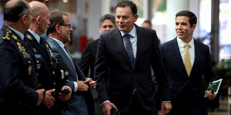 Un gruppo di uomini in abiti formali, compresi abiti e uniformi militari, camminano insieme e conversano sul nuovo piano immigrazione portogallo. Alcuni hanno in mano documenti e sembrano trovarsi in un ambiente professionale o ufficiale. Lo sfondo mostra un ambiente interno con persone e un arredamento moderno.