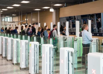 Un gruppo di turisti britannici è in fila al checkpoint di sicurezza EES. Ogni persona entra da un cancello alla volta. L'area è un aeroporto dal design moderno con un soffitto alto e un'illuminazione ampia e luminosa.