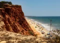Una vista panoramica della Praia da Falesia in Algarve, meta preferita dalle vacanze dei portoghesi, che si mostra con alte scogliere rossastre sulla sinistra con una spiaggia sabbiosa che si estende lungo le calme onde dell'oceano. Diverse persone si rilassano sulla spiaggia con ombrelloni colorati e asciugamani sparsi qua e là.