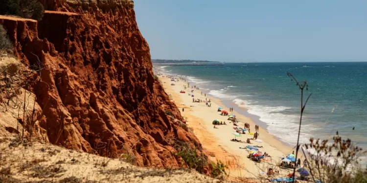 Una vista panoramica della Praia da Falesia in Algarve, meta preferita dalle vacanze dei portoghesi, che si mostra con alte scogliere rossastre sulla sinistra con una spiaggia sabbiosa che si estende lungo le calme onde dell'oceano. Diverse persone si rilassano sulla spiaggia con ombrelloni colorati e asciugamani sparsi qua e là.