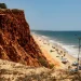 Una vista panoramica della Praia da Falesia in Algarve, meta preferita dalle vacanze dei portoghesi, che si mostra con alte scogliere rossastre sulla sinistra con una spiaggia sabbiosa che si estende lungo le calme onde dell'oceano. Diverse persone si rilassano sulla spiaggia con ombrelloni colorati e asciugamani sparsi qua e là.