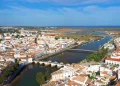 Veduta aerea di Tavira caratterizzata da un mix di casa bianche tipiche dell'Algarve da vendere. Il fiume Gilao divide la città con numerosi ponti che la attraversano, conducendo a una costa panoramica e a una zona paludosa in lontananza sotto un cielo azzurro e limpido.