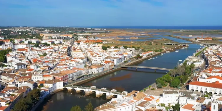 Veduta aerea di Tavira caratterizzata da un mix di casa bianche tipiche dell'Algarve da vendere. Il fiume Gilao divide la città con numerosi ponti che la attraversano, conducendo a una costa panoramica e a una zona paludosa in lontananza sotto un cielo azzurro e limpido.