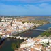 Veduta aerea di Tavira caratterizzata da un mix di casa bianche tipiche dell'Algarve da vendere. Il fiume Gilao divide la città con numerosi ponti che la attraversano, conducendo a una costa panoramica e a una zona paludosa in lontananza sotto un cielo azzurro e limpido.