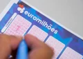 Primo piano di un biglietto della lotteria della vincita record Euromilhoes compilato da una mano che tiene una penna a Porto. Il biglietto presenta diverse griglie numerate in cui il giocatore può contrassegnare i numeri scelti. Il logo e il marchio Euromilhões sono visibili in alto.