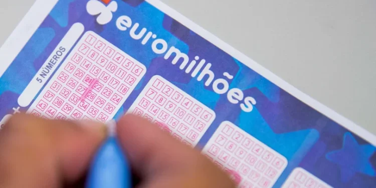 Primo piano di un biglietto della lotteria della vincita record Euromilhoes compilato da una mano che tiene una penna a Porto. Il biglietto presenta diverse griglie numerate in cui il giocatore può contrassegnare i numeri scelti. Il logo e il marchio Euromilhões sono visibili in alto.