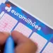 Primo piano di un biglietto della lotteria della vincita record Euromilhoes compilato da una mano che tiene una penna a Porto. Il biglietto presenta diverse griglie numerate in cui il giocatore può contrassegnare i numeri scelti. Il logo e il marchio Euromilhões sono visibili in alto.