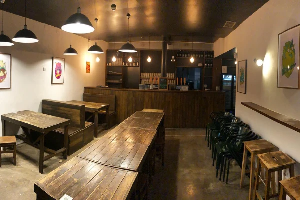 Un interno del Duque Brewpub accogliente con tavoli e panche in legno, che ricorda le migliori birrerie. La stanza poco illuminata ha lampade a sospensione appese al soffitto. Sul retro del bancone del bar sono presenti numerose bottiglie e rubinetti. Sedie verdi, sgabelli e opere d'arte incorniciate sono situati lungo le pareti.