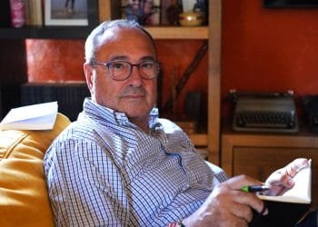 Enrico Berardi, manager e scrittore, con gli occhiali e una camicia a quadri, è seduto su una sedia gialla, guardando la telecamera. Tiene una penna e un taccuino aperto. Dietro di lui c'è una libreria con libri, foto incorniciate e una macchina da scrivere.