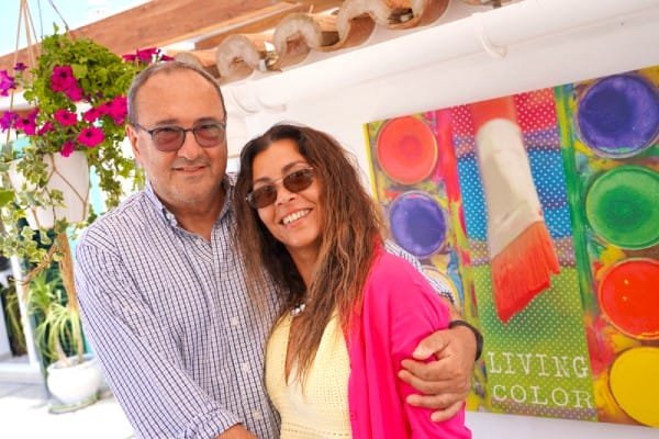 Enrico Berardi, manager e scrittore, e la moglie Naiby Martinez sorridenti e abbracciati in un ambiente luminoso e colorato. L'uomo porta gli occhiali e una camicia a quadretti blu. La donna ha i capelli lunghi, occhiali da sole e una giacca rosa. Dietro di loro c'è un dipinto vibrante di un pennello e una tavolozza di colori.