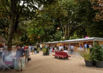 Tutti gli Eventi con Esposizioni e Mostre della Fiera del Libro di Porto all'aperto sotto una tettoia di alberi presenta varie bancarelle di venditori. Le persone camminano, chiacchierano e navigano. Una prominente panchina rossa e grandi piante in vaso decorano lo spazio. Le luci delle corde pendono sopra, aggiungendo un'atmosfera calda alla scena degli eventi serali.