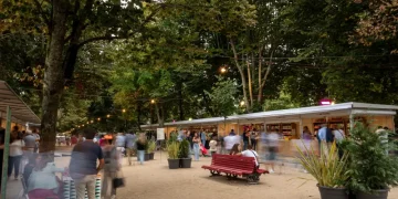 Tutti gli Eventi con Esposizioni e Mostre della Fiera del Libro di Porto all'aperto sotto una tettoia di alberi presenta varie bancarelle di venditori. Le persone camminano, chiacchierano e navigano. Una prominente panchina rossa e grandi piante in vaso decorano lo spazio. Le luci delle corde pendono sopra, aggiungendo un'atmosfera calda alla scena degli eventi serali.