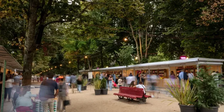 Tutti gli Eventi con Esposizioni e Mostre della Fiera del Libro di Porto all'aperto sotto una tettoia di alberi presenta varie bancarelle di venditori. Le persone camminano, chiacchierano e navigano. Una prominente panchina rossa e grandi piante in vaso decorano lo spazio. Le luci delle corde pendono sopra, aggiungendo un'atmosfera calda alla scena degli eventi serali.