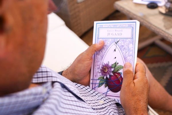 Persona con in mano un libro intitolato "Jugaad" di Enrico Berardi con un'illustrazione floreale e un cuore in copertina. La persona indossa una camicia a quadretti bianchi e blu ed è seduta in quello che sembra essere un soggiorno o un'area salotto, con mobili in vimini e un tappeto a motivi geometrici sullo sfondo.