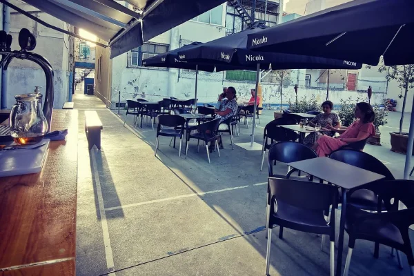 Un'invitante area salotto della Maria Piolas Dehors a Lisbona all'aperto presenta diversi tavoli e sedie neri sotto grandi ombrelloni neri. Alcuni avventori sono seduti e conversano, mentre la luce del sole proietta lunghe ombre sul pavimento di cemento. A sinistra, un bancone con una caffettiera.