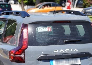 Il veicolo, probabilmente guidato da un autista TVDE, è una Dacia grigia raffigurata da dietro, con un adesivo TVDE sul lunotto posteriore. È ambientato in una location all'aperto in Portogallo con altre auto visibili sullo sfondo.