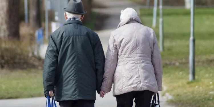 Una coppia di anziani centenari cammina mano nella mano lungo un sentiero a Lisbona in Portogallo, portando ciascuno una borsa. L'uomo indossa una giacca e un cappello scuri, mentre la donna indossa un cappotto rosa chiaro. Il percorso è fiancheggiato da erba e alberi, con lampioni lungo il percorso.